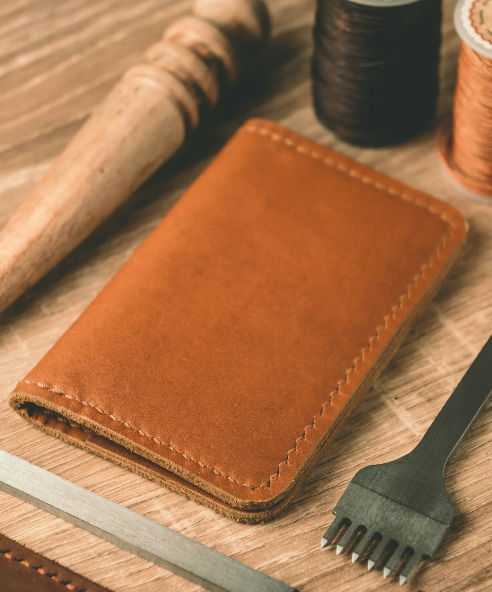 A hand-stitched tan leather holder on a workbench beside leather tools and spools of waxed thread